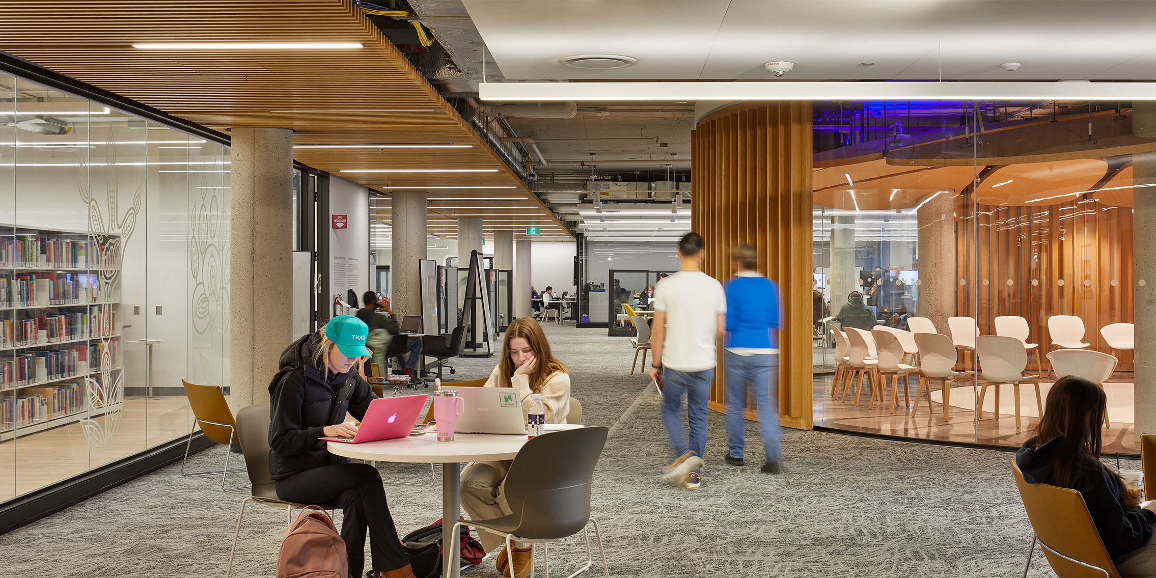 Students studying at tables in a bright, open library with bookshelves and the Kalihwíy̲o̲ Circle in the background. 