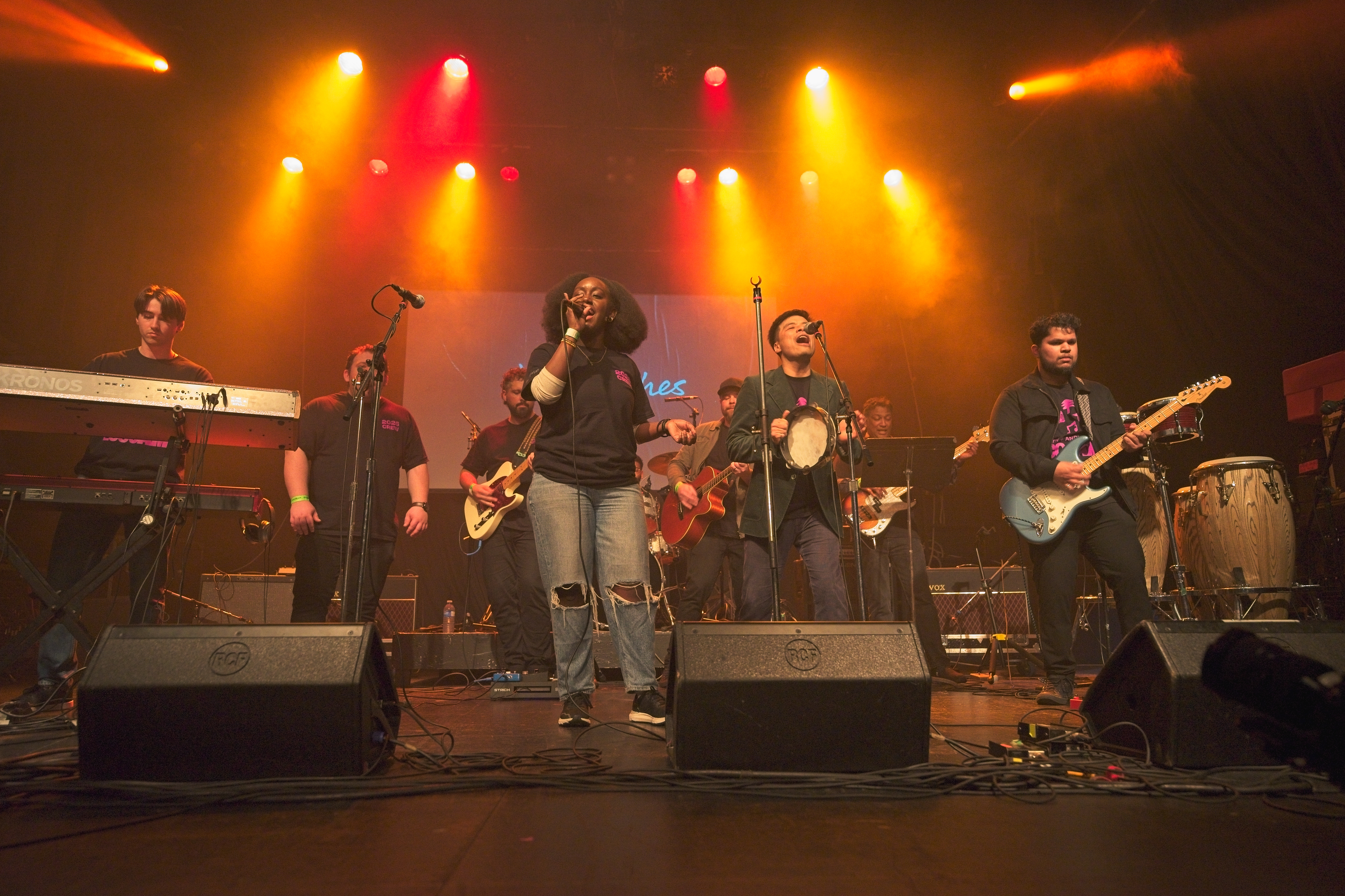 A full band performs on stage under orange and yellow lights, with microphones, guitars, keyboards, and drums visible.