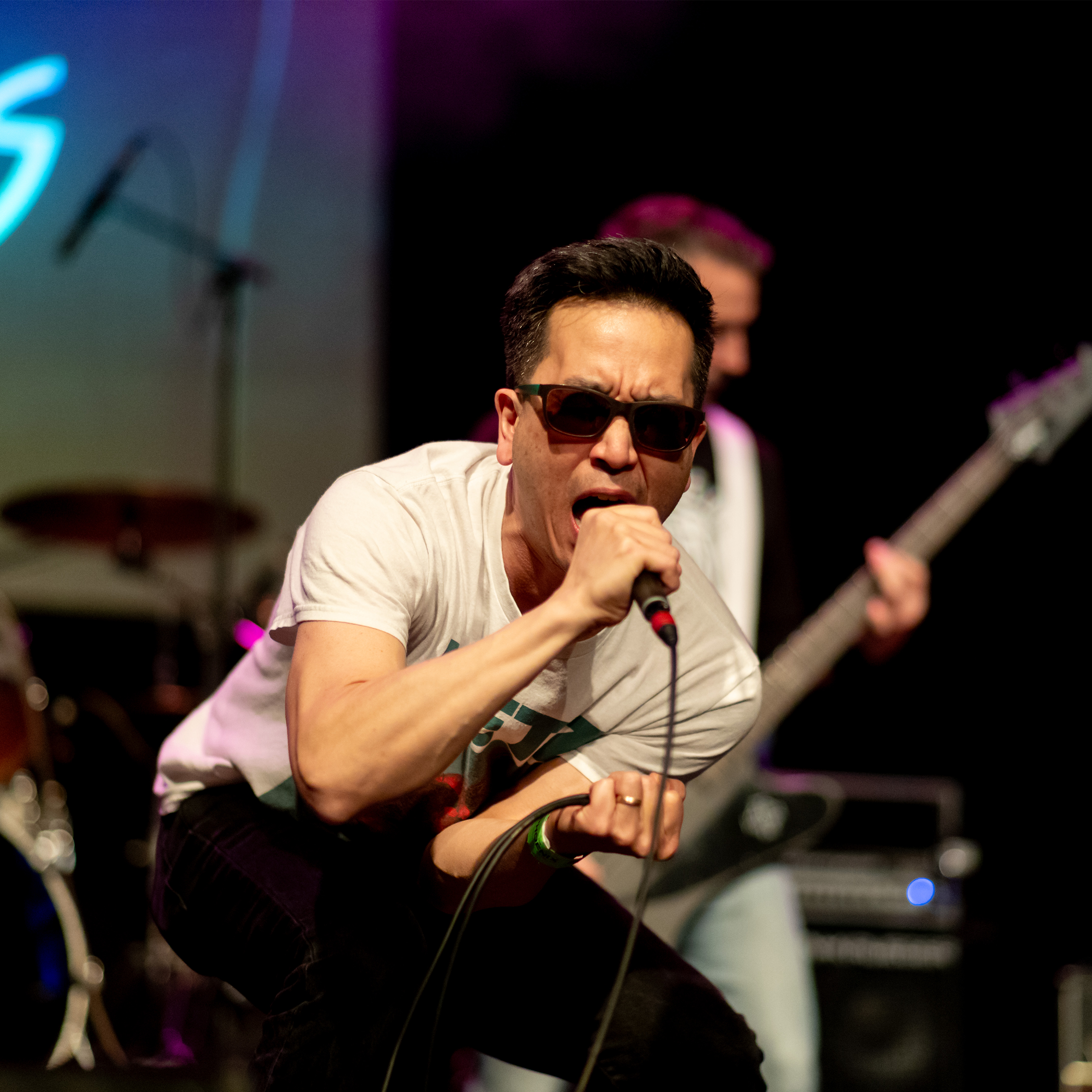 A performer crouches at the front of the stage singing into a handheld microphone, with band members and instruments behind.