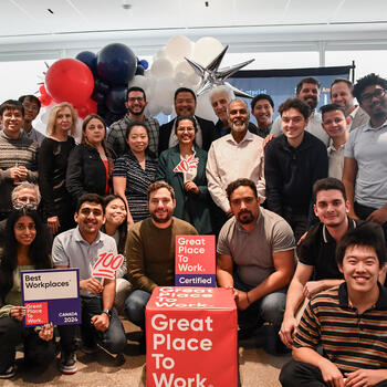 A group photos of employees around a red cute with the white text reading "Great Place to Work" and award plaques.