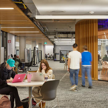 Students studying at tables in a bright, open library with bookshelves and the Kalihwíy̲o̲ Circle in the background. 