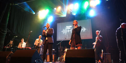 Two singers perform on stage in the foreground, with the rest of the band in the background. "ThunderTRUSS" sign appears behind.