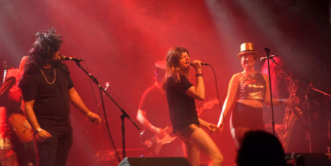 Band performs on stage. Three singers wear costumes and wigs.