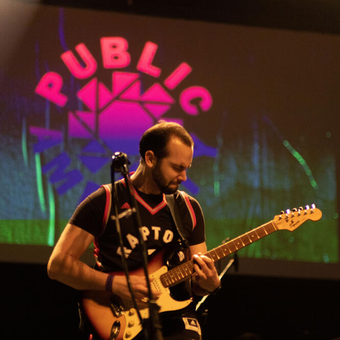 Guitarist playing on stage wearing Raptors jersey and standing in front of screen that says "Public Amenity"