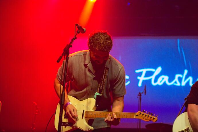 A man plays an electric guitar and sings into a microphone on stage under red and yellow lighting, with the word "Flash" visible in the background..